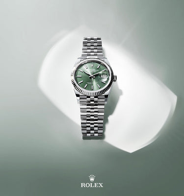 Silver watch with green face on a white platform against a gray background, featuring the Rolex logo.